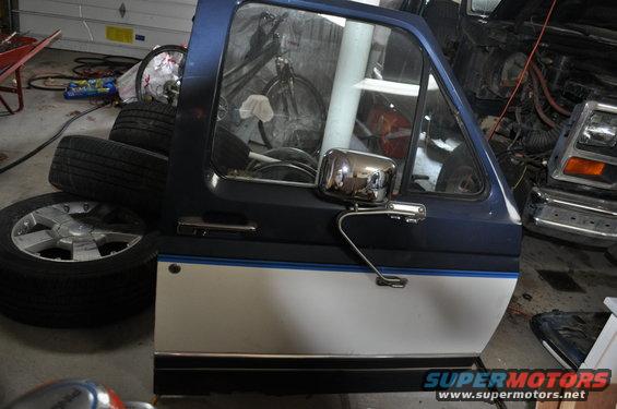Blue and white Ford Bronco door with chrome mirror, stacked tires nearby, another vehicle in background