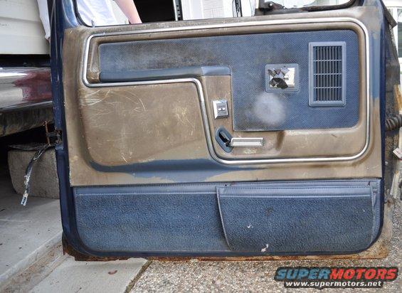 Interior door panel with brown and blue fabric, showing a window crank, door handle, and air vent