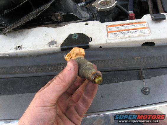 Hand holding a dirty fuel injector with yellow top, in front of a white vehicle's engine bay