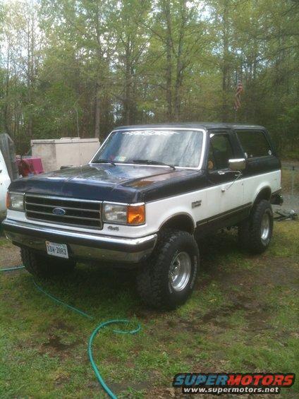 Two-tone Ford Bronco parked on grass with trees in background. License plate visible