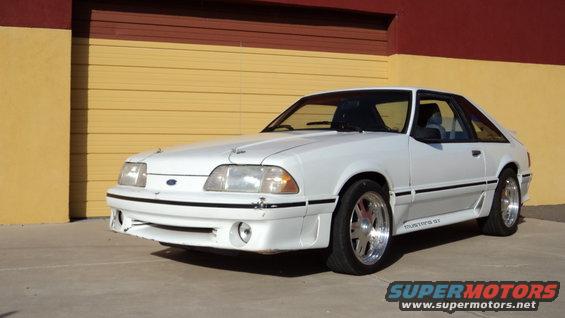 White Ford Mustang GT parked in front of a yellow and red garage door. It has chrome wheels and a black stripe along the side