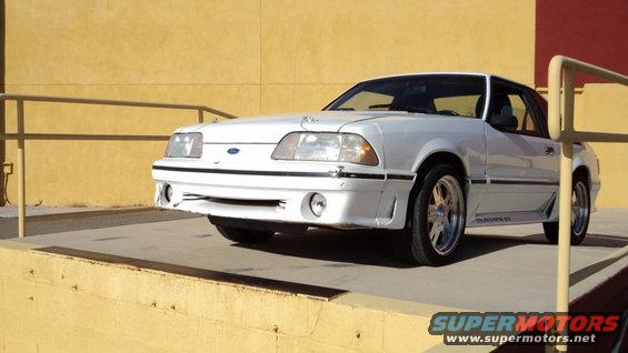 White Ford Mustang with a damaged front bumper, parked on a concrete surface next to a yellow wall
