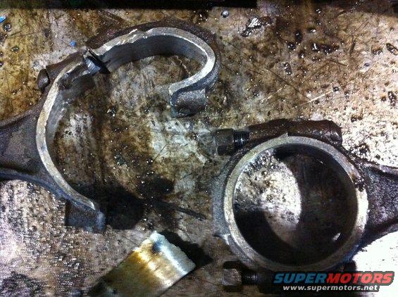 Two broken engine connecting rods on a dirty workbench. One rod is in two pieces, the other is cracked