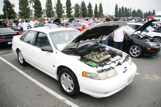 White sedan with hood open, showing engine. Parked at an outdoor car show