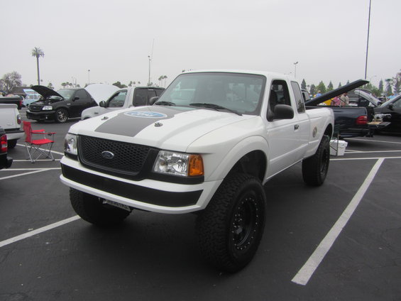 White Ford Ranger pickup with black racing stripes on hood, lifted suspension, and off-road tires. Other vehicles are parked nearby