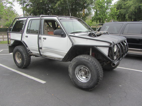 White SUV heavily modified with a full roll cage, off-road tires, and custom front end. Another black SUV is parked behind it
