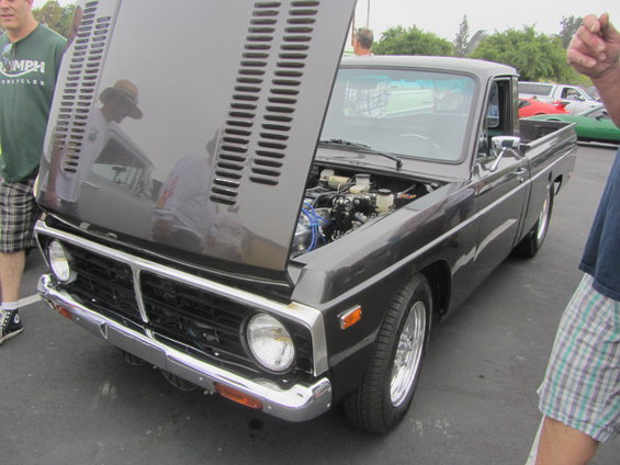 Dark gray pickup truck, hood open, showing engine. Chrome grille and bumper. Chrome wheels. Visible in background: red and green cars