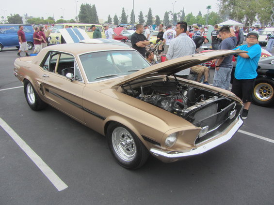 Gold 1967 Ford Mustang coupe with hood open, showing engine. Chrome wheels, black side trim