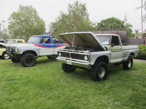 Two lifted pickup trucks parked on grass. One is blue and white with red stripes, the other is light green and silver with its hood open