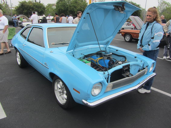 Light blue coupe with engine exposed, woman looking at it. Other cars and people in background