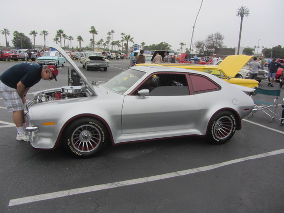 Silver 1974 Ford Pinto coupe with red trim, hood open, guy looking at engine. Other cars in background