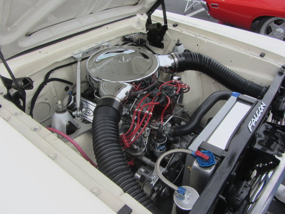 Engine bay of a white car, showing a V8 engine with chrome air cleaner and red spark plug wires. The word "FALCON" is visible on a radiator support