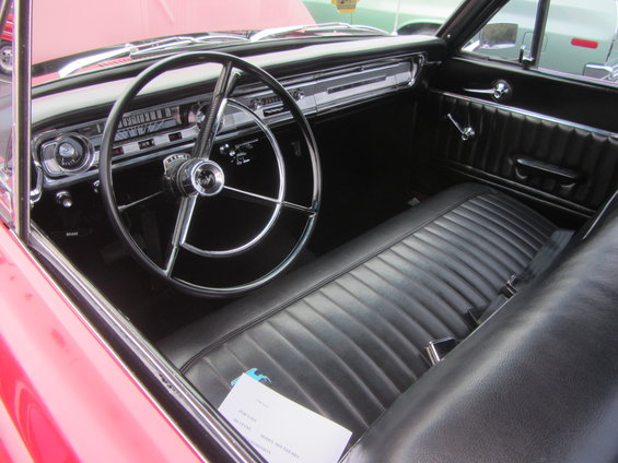 Red 1964 Ford Ranchero interior with black vinyl bench seat, dashboard, and steering wheel. A document is on the seat