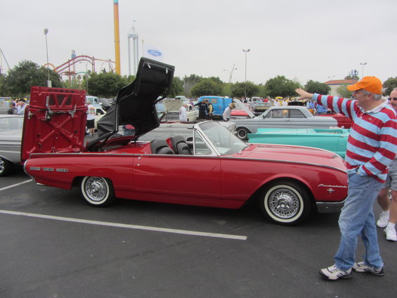 Red convertible with its top mechanism open, showing a man pointing. Other cars and amusement park rides are in the background