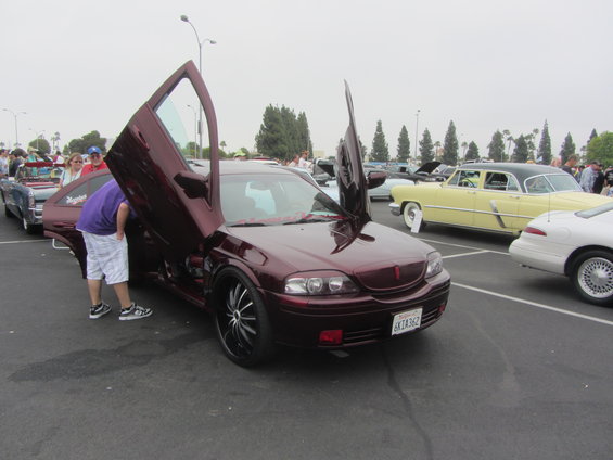 Maroon sedan with gullwing doors open, people looking at it, other cars in background