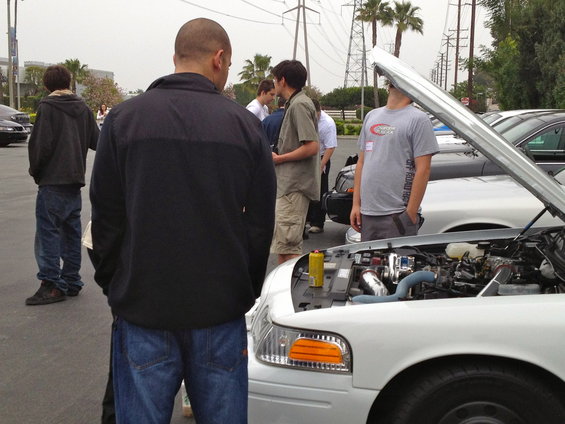 White Ford Crown Victoria, hood open showing engine bay. Several people stand around it