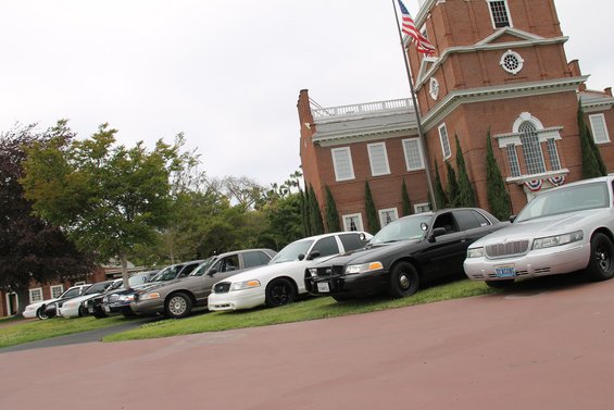 Several Ford Crown Victoria police cars parked in a row in front of a brick building with an American flag