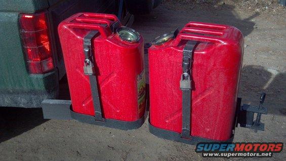 Two red jerry cans attached to a vehicle's rear bumper. The cans are secured with black straps