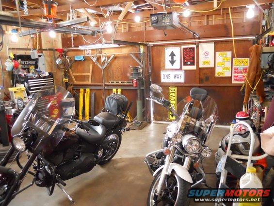 Two motorcycles parked inside a cluttered garage with tools and signs on the wall