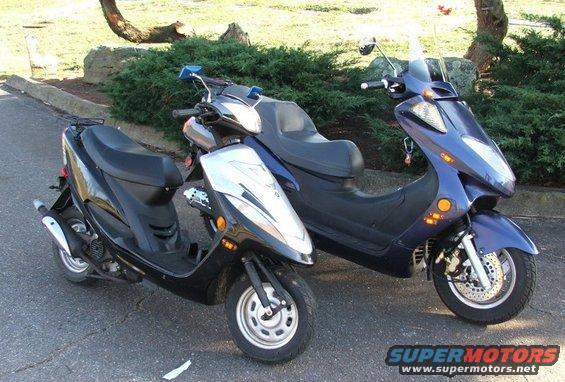 Two scooters parked side-by-side on an asphalt surface with a grassy area and trees in the background