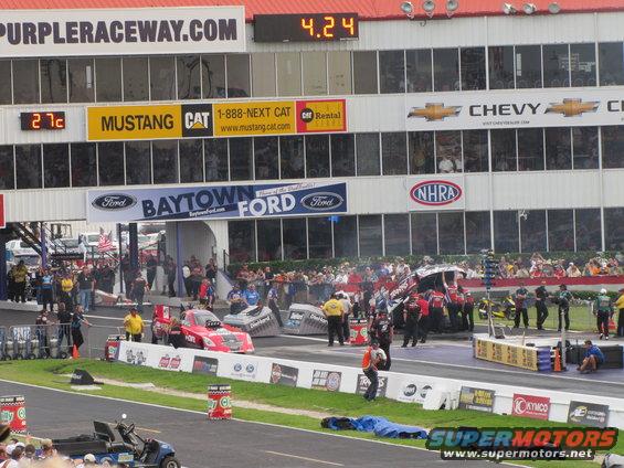 Drag strip with race cars lined up, crowd watching, and scoreboards overhead
