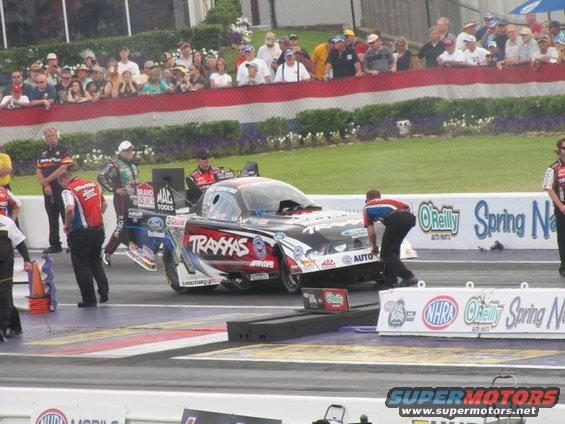 Drag racing car with 'Traxxas' livery being pushed by crew members on a track, spectators in background