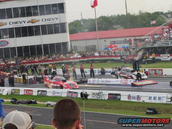 Two drag racing cars on a track, one red and one black with red accents, with spectators in the background