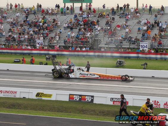 Drag racing car with 'Parts Plus' livery on track, spectators in stands