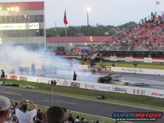 Drag strip with smoke, spectators in bleachers, and a blurred race car in motion