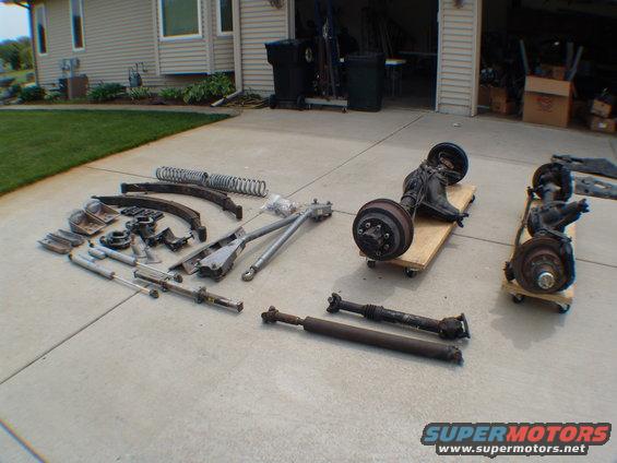 Assorted vehicle suspension and drivetrain parts laid out on a concrete driveway, including axles, springs, shocks, and driveshafts