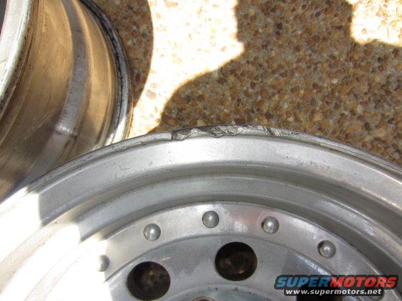 Close-up of a damaged aluminum wheel rim with visible dents and scratches on the lip. Another wheel is partially visible in the background