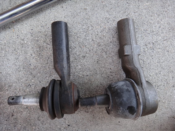 Two tie rod ends, one with a silver threaded stud and black rubber boot, the other with a dark metal body and black rubber boot