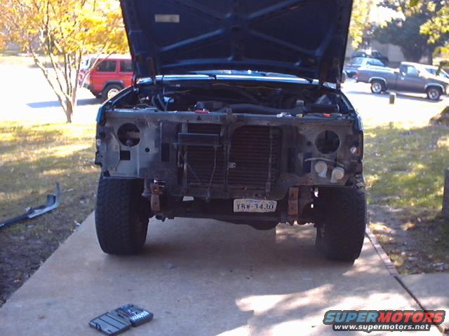 Front of a dark blue Ford truck with hood up, grille and bumper removed. License plate TRW-3430 visible. Red SUV and gray pickup in background