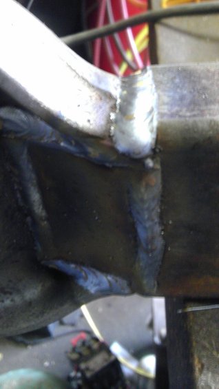 Close-up of a metal part with visible welds. Some red and yellow material is out of focus in the background