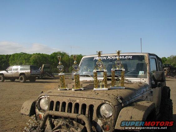 Mud-covered Jeep Wrangler with trophies on hood, other muddy vehicles in background