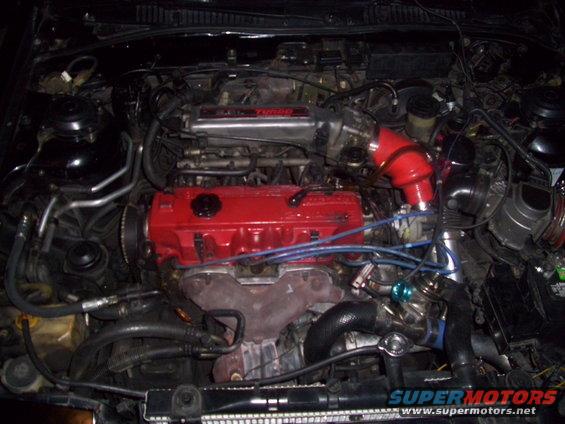 Engine bay with a red valve cover, turbocharger, and red intake piping. Text on the bottom reads 'SUPERMOTORS'