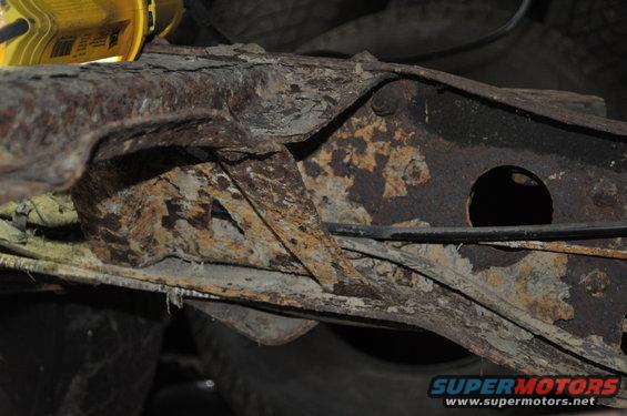 Close-up of heavily rusted metal frame components, possibly from a vehicle's undercarriage. A yellow work light is visible at the top left