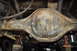 1986 Ford Bronco - The underside of a vehicle's rear differential shows significant rust and wear, with brake lines and suspension parts attached.