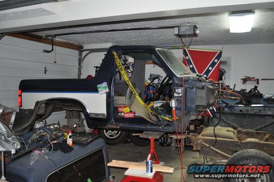 Disassembled blue and white Ford Bronco in garage, front end removed, on jack stands