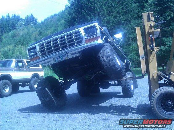 Black 1978 Ford Bronco lifted with large tires, being moved by a yellow forklift. Another Bronco is parked behind it