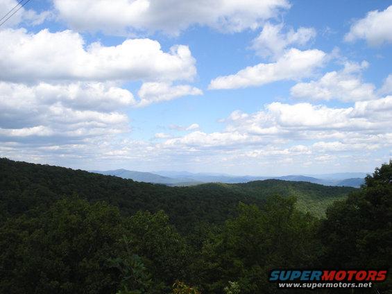 Mountain landscape with green trees and blue sky with clouds. Watermark reads SUPERMOTORS