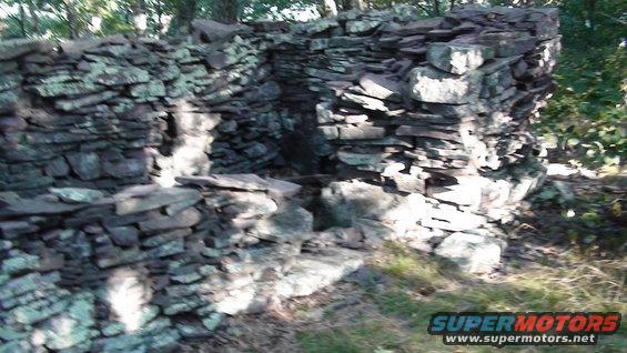 Stone structure in wooded area, partially collapsed, with green moss on stones