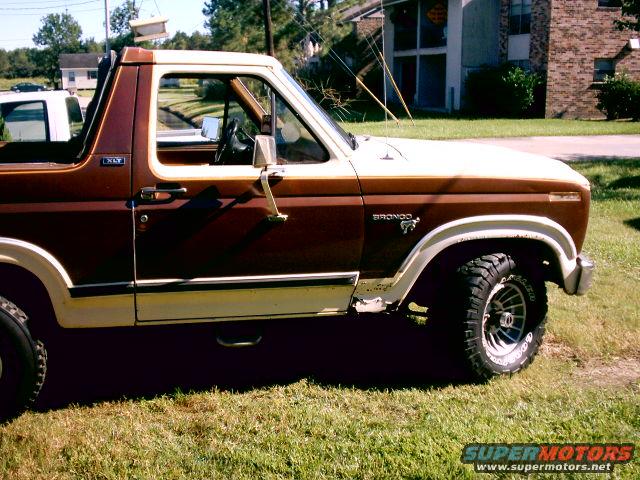 Brown and white 1980 Ford Bronco side view, grass, buildings in background