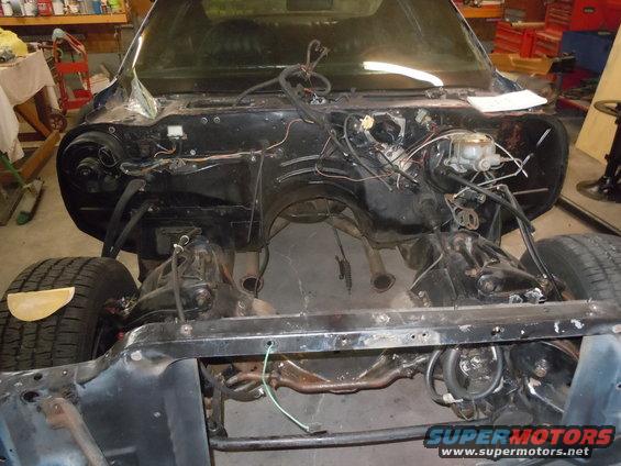 Disassembled car engine bay, dark blue exterior, front view, showing engine mounts and exhaust pipes