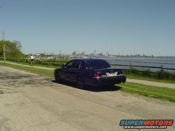 Dark sedan parked on a road next to a body of water with a city skyline in the distance