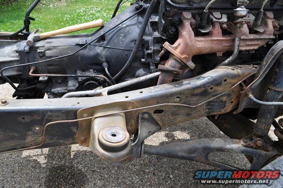 Close-up of vehicle frame with engine block, transmission, and exhaust manifold visible. A hammer rests on the transmission