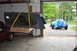 1986 Ford Bronco - A dark blue camper shell is suspended by yellow straps, with a red car visible below and another vehicle covered in a blue tarp in the back…