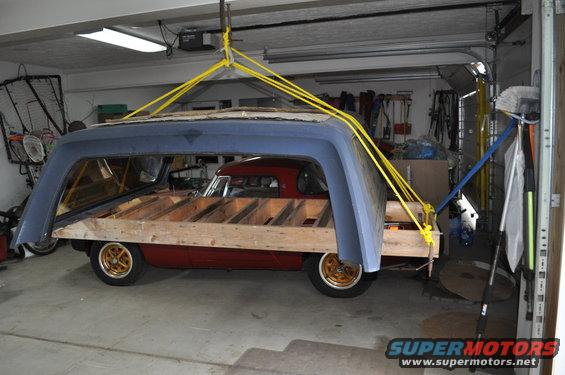 Red car body with wooden frame and blue camper shell being lifted by yellow straps in a garage