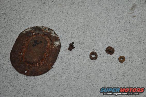 Rusty circular plate with faint writing, a screw, and three nuts on a gray surface