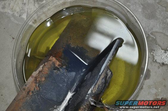 Rusted metal part soaking in yellow liquid, with Supermotors.net logo visible